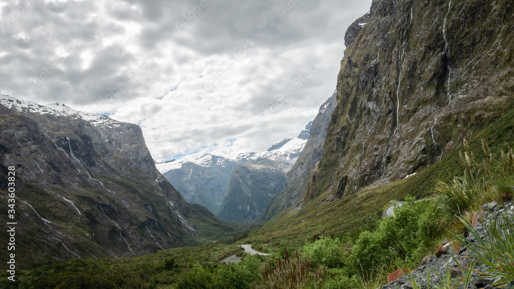 Green valley full of waterfalls and snowy mountains in backdrop shot on overcast day. Photo taken in Fiordland National Park, New Zealand