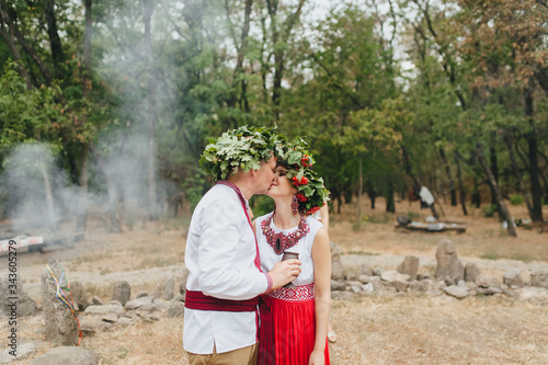 The bride and groom in the Ukrainian traditions of paganism with wreaths on their heads and embroidered clothes kiss and hug in nature in the forest at a ceremony. Photography, concept.