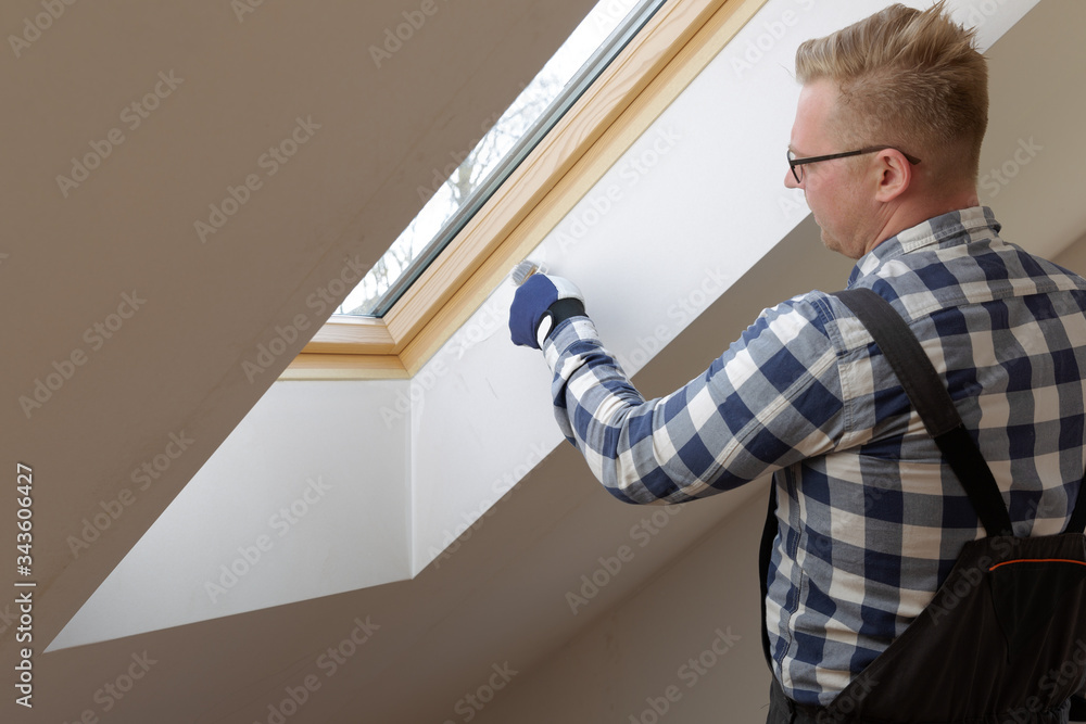 Home improvement concept, handyman painting a wall with a white paint near roof window in attic.