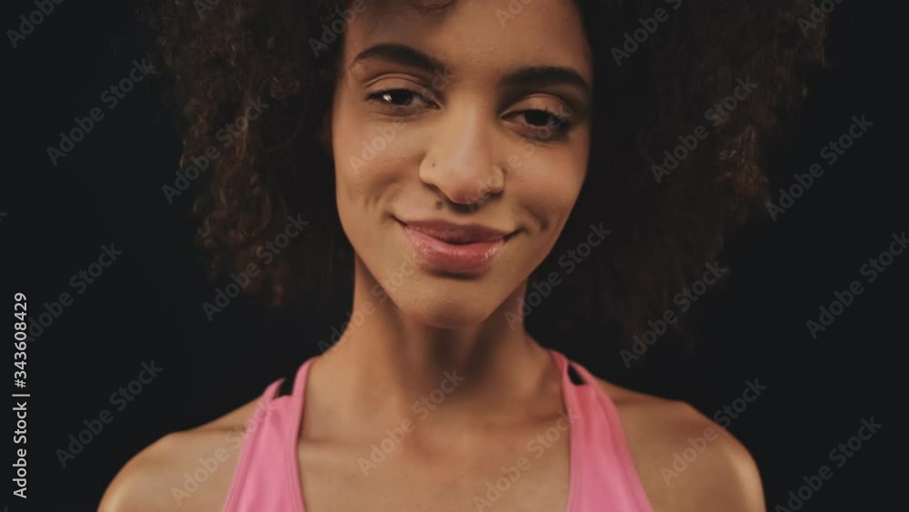 A close-up view of a pleased athletic african american woman in sportswear is smiling and looking to the camera isolated over black background A close-up view of a pleased athletic african american woman in sportswear is smiling and looking to the camera isolated over black background