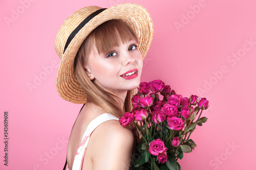 Beauty blonde woman in straw hat with fresh flowers on pink background.