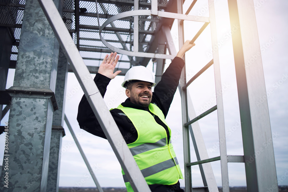 Industrial factory construction worker waving and climbing metal ladder of production plant in sunset.