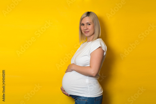a happy pregnant woman in a white t shirt and jeans stands sideways on a yellow background