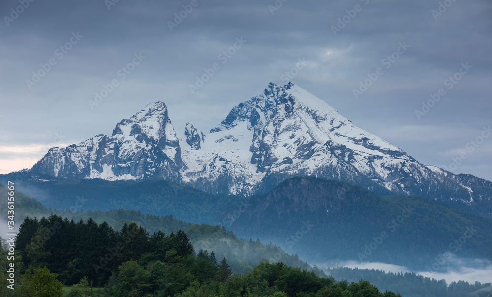 Snow covered mountain peaks of Watzmann at Berchtesgaden with clouds and morning fog, green forest in foreground, Bavaria.