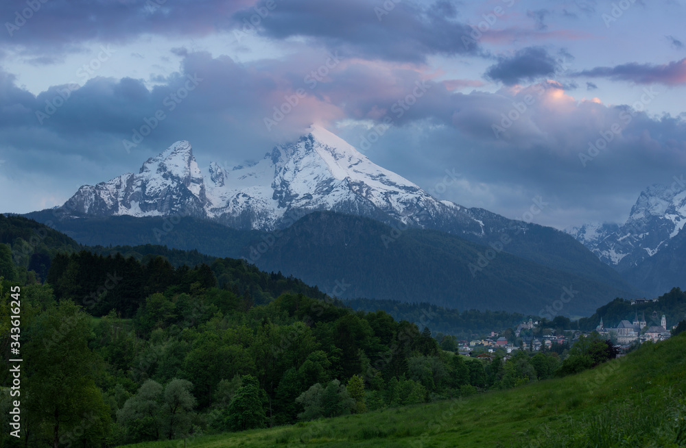 Snow covered mountain peaks of Watzmann and city Berchtesgaden with clouds during sunset, green forest and fields in foreground, Bavaria.