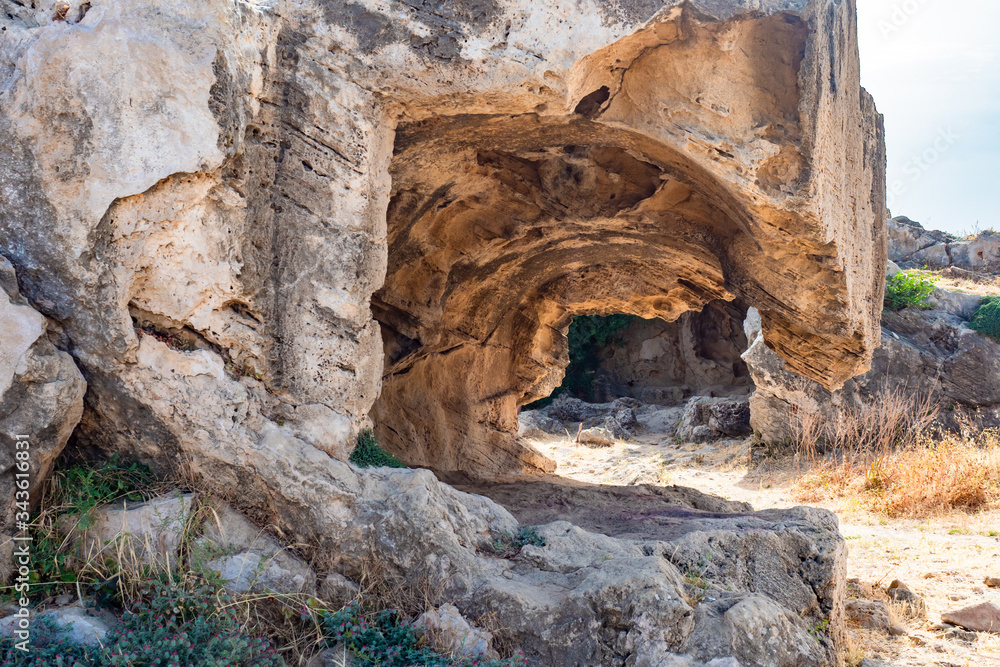 A rocky cave against a background of sparse vegetation. Sights Of Cyprus. Archaeological Park of the city of Paphos. Paphos open-air Museum. Travel to the Mediterranean.