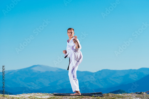 Blond karate athlete does kata on top of a mountain while performing a line up of kicks, punches and blocks on top of a mountain on a sunny day.