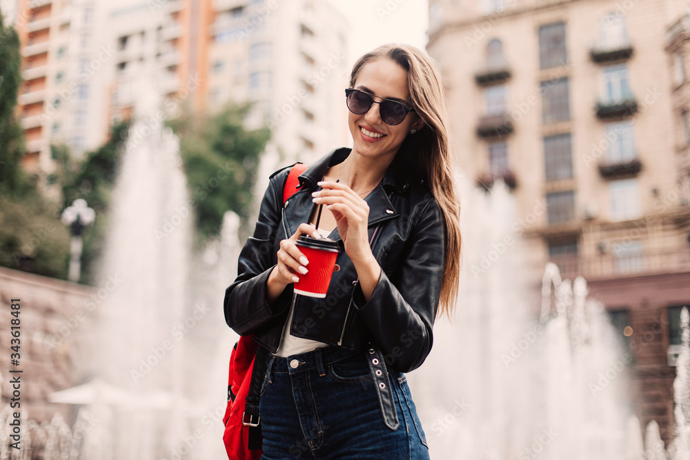 young attractive stylish smiling woman having fun in city, positive, emotional, long hair, wearing black jacket, fashionable jeans. holds hands takeaway coffee.