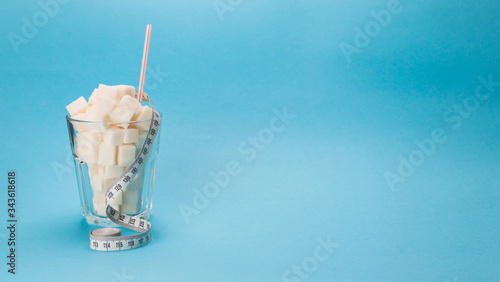 Glass cup with a straw full of cubes of white sugar on a blue background. A measuring tape is wrapped around the glass.