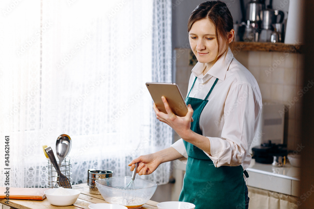 Fototapeta premium Woman looking to laptop and reading recipe for cooking dishes. Cooking at home concept, lifestyle. Preparing the dough, baking in the kitchen.