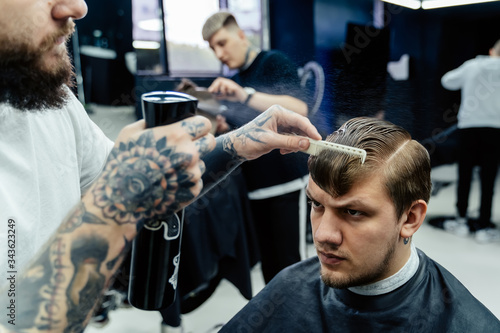 Attractive male is getting a modern haircut in barber shop. Tattooed Barber wets hair by spray and combs them.