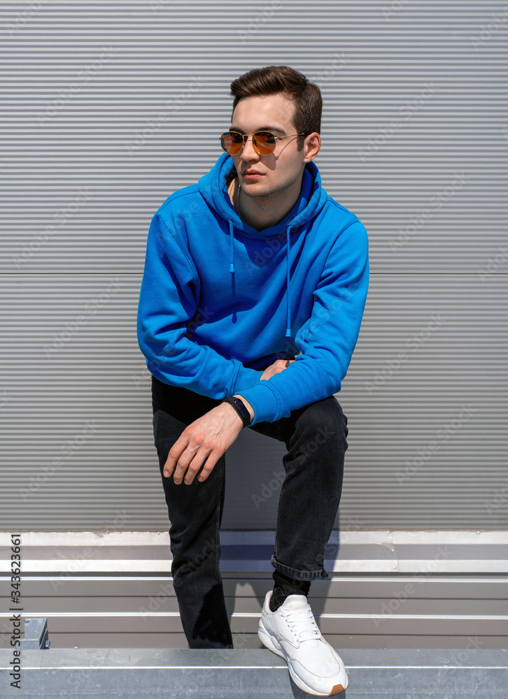 slender young man in red sunglasses, with an earring, dressed in a blue  hoodie, black jeans, white sneakers and black fitness tracker, standing  against a gray textured wall in the afternoon Stock