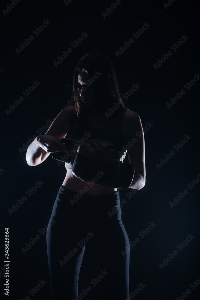 Silhouette portrait with dark contrast of a young fitness girl putting on her boxing gloves