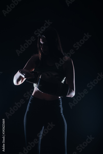 Silhouette portrait with dark contrast of a young fitness girl putting on her boxing gloves