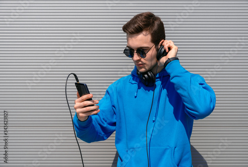 student in black round glasses, holds smartphone in his right hand and headphones in his left hand, wearing blue hoodie and black fitness tracker, listening music against a gray wall in the afternoon