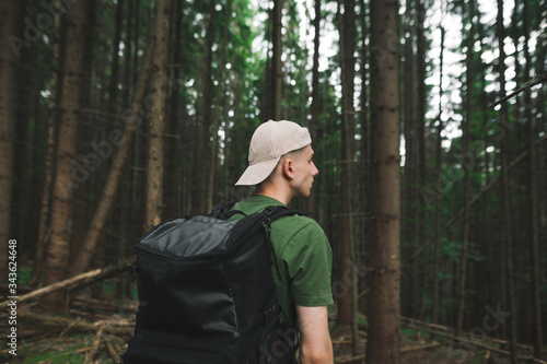 The back of a young man in casual clothes and with a backpack walks through the pristine coniferous forest and looks away. Guy tourist walks through the forest.