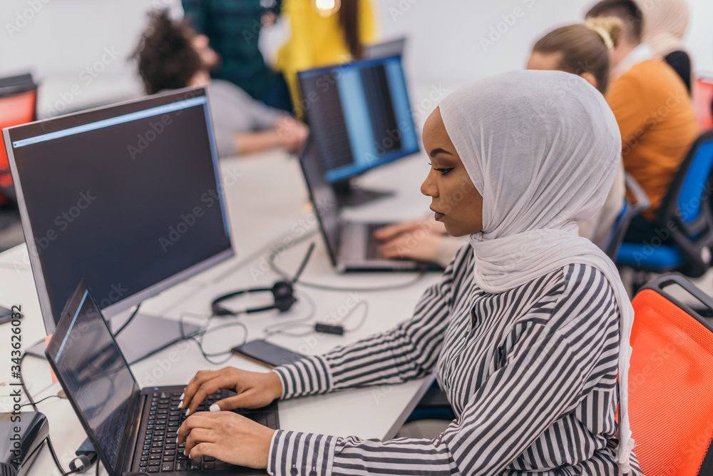 Picture from behind of a female employee with white hijab sitting at her desk and working on her laptop Picture from behind of a female employee with white hijab sitting at her desk and working on her laptop
