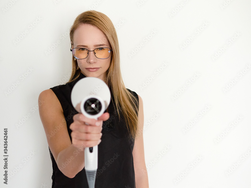Fototapeta premium Confident blonde female hairdresser in glasses and black apron standing against white wall in trendy barbershop salon holding professional hair dryer