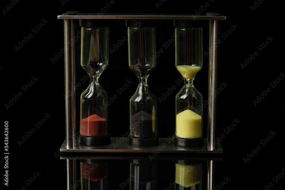 Fototapeta premium Three hourglasses with colorful sand stand on a black glossy glass, in which their reflection is visible.
