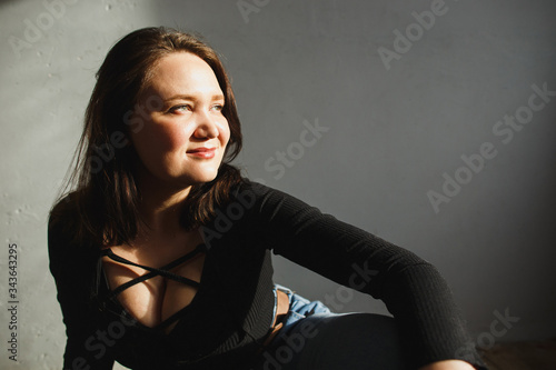 Young woman sits on the floor of the room and provocative looks to the sunlight. She clothed in black body with deep neckline and blue jeans.