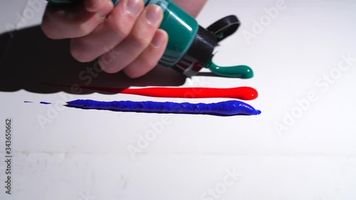 This closeup video shows an anonymous hand squeezing green paint from a tube onto a  white surface, next to other colors of paint. photo