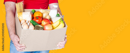 Grocery delivery courier man in red uniform holds cardboard box with fresh vegetables, fruits and other food on orange background. Express food delivery, donation concept.