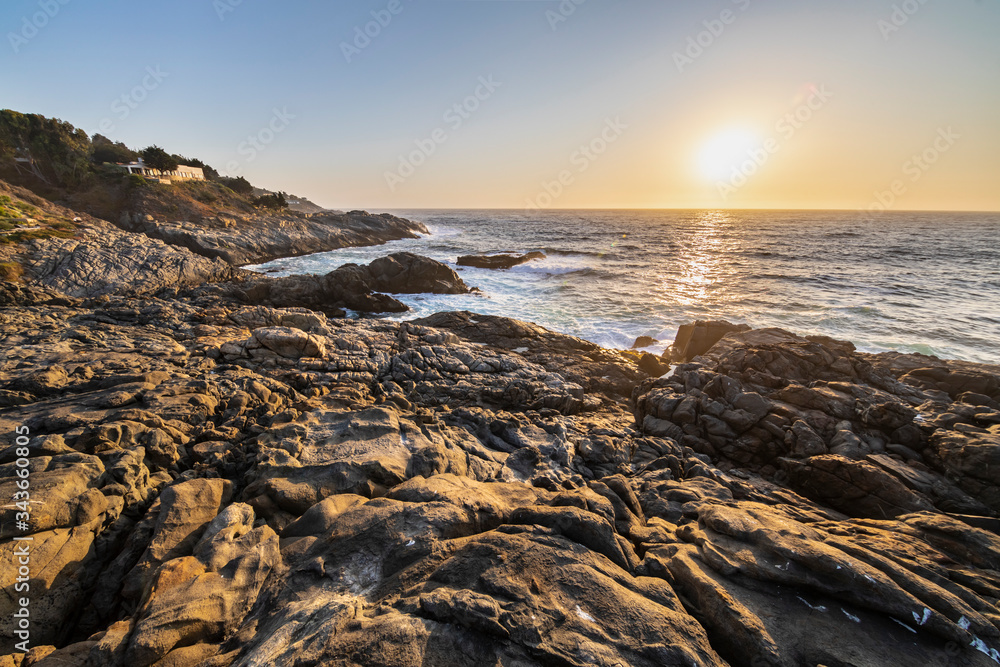 An amazing view of the sunset over the water in the Chilean coast. An idyllic beach scenery with the sunlight illuminating the rocks with orange tones and the sea in the background under a moody sky
