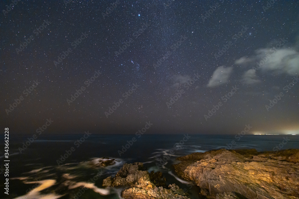 A view of the Milky Way and the stars at a starry night above the sea in the Pacific Ocean with the waves crashing the rock cliffs illuminated by the surrounding houses making an idyllic nature scene