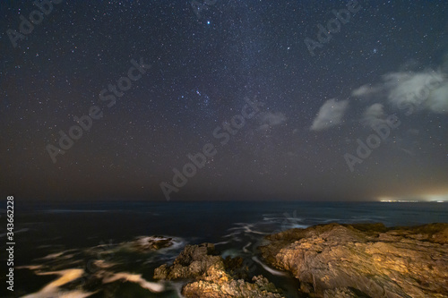 A view of the Milky Way and the stars at a starry night above the sea in the Pacific Ocean with the waves crashing the rock cliffs illuminated by the surrounding houses making an idyllic nature scene