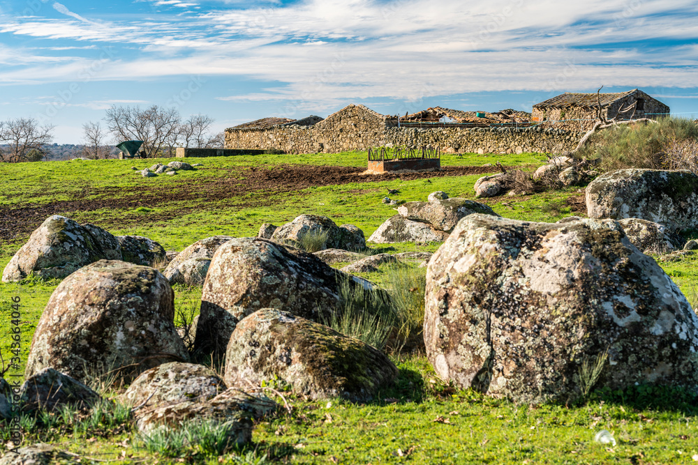 Farmland inside the Spanish rural countryside. Rock boulders, green pasture, agriculture shelter and a warehouse conform the old farm with a granite construction. A view during a cloudy winter day. 