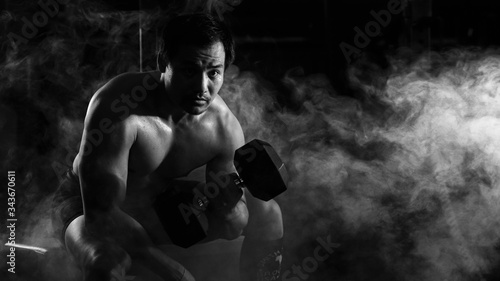 asian athletic strong man sitting and having weight lifting with dumbbells with one arm in gym and fitness center with smoke and dark background in black and white