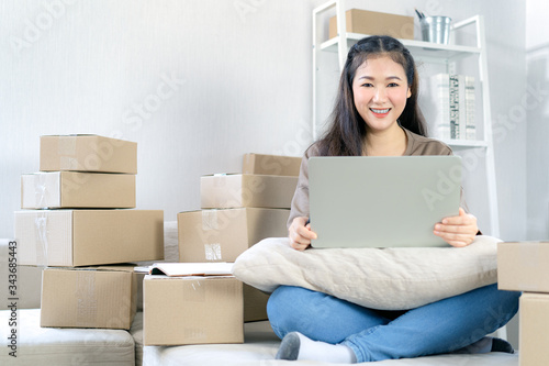 Young asian attractive smiling girl entrepreneur work at home and use her laptop for doing business and check customer data. Packaging parcels boxes in background. Entrepreneur activity concept.