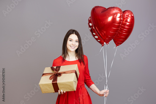 Joyful girl with balloons and a gift. A teenager girl in a red dress on a gray background holds balloons and a gift box in her hands. © ALEXEY