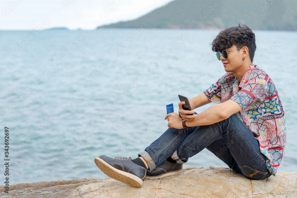 Smiling young smart Asian man guy sitting on rocky seacoast beach using smartphone with internet for online shopping and credit card for e-banking payment. E-commerce and wireless technology concept