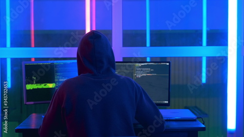 Back view of hacker in a black jacket and a hood on his head writing hacker programs. He working on personal computer in hacker dark room.