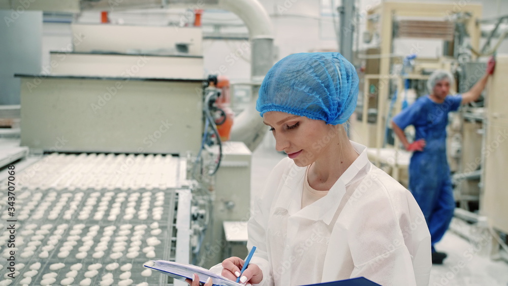 Candy factory. Controller checking conveyor with candies. Young woman in uniform holding folder and inspecting conveyor belt with fresh candies in confectionery factory. Candy factory. Controller checking conveyor with candies. Young woman in uniform holding folder and inspecting conveyor belt with fresh candies in confectionery factory.