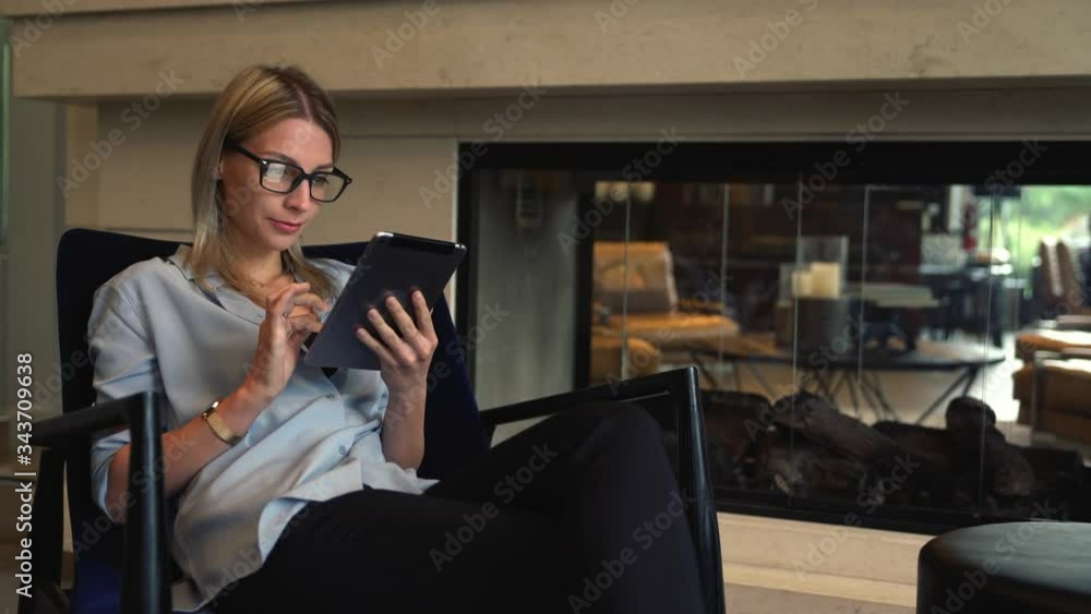 Caucasian attractive young woman in optical eyeglasses sitting in comfortable chair of hotel lobby interior and enjoying free time for communicate in social network via application on digital tablet
