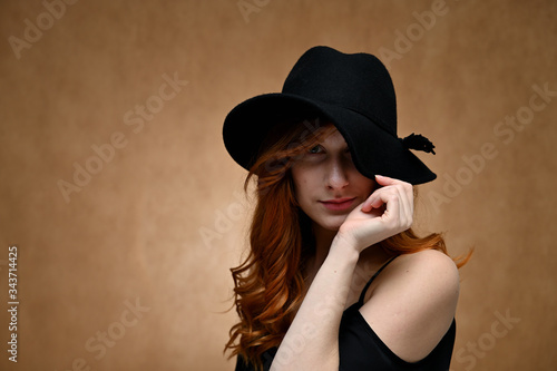 Model posing in a black hat. Portrait of a young pretty woman with long red hair on a beige background.