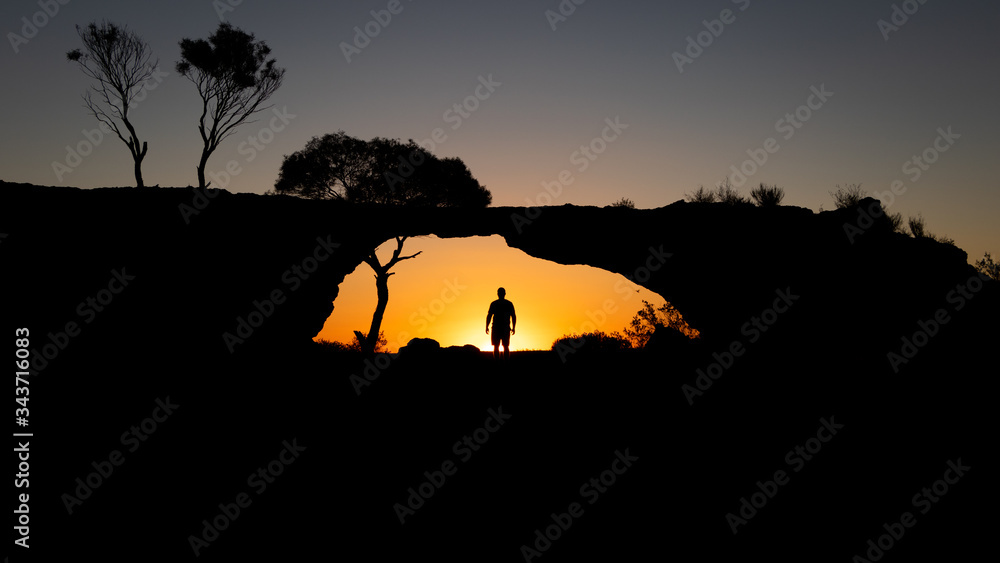 Silhouette of a man standing under a rock bridge at sunset