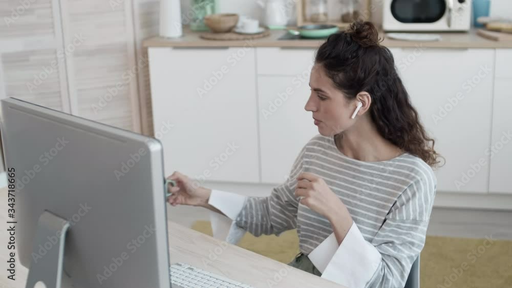 Caucasian woman wearing wireless earphones sitting at table at PC, communicating by video link and drinking tea. Then she putting food into microwave and continuing chat Caucasian woman wearing wireless earphones sitting at table at PC, communicating by video link and drinking tea. Then she putting food into microwave and continuing chat