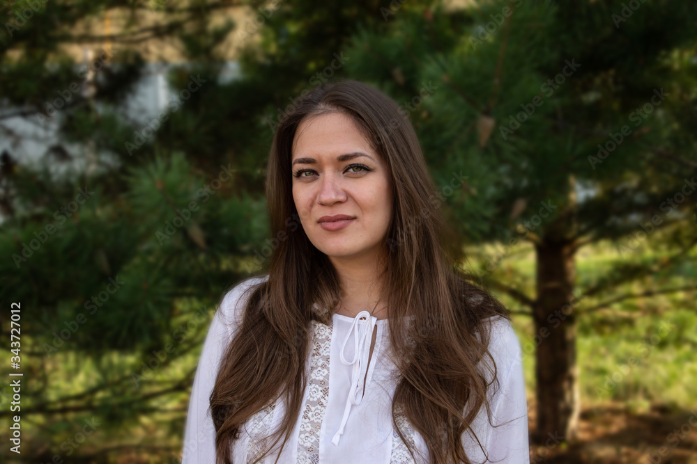 Portrait of a young brown-haired beautiful girl with green eyes and a white blouse on a background of trees Portrait of a young brown-haired beautiful girl with green eyes and a white blouse on a background of trees