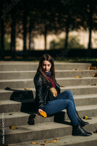Stylish beautiful lady in a black leather jacket with a black bag and sweatshirt. Female sits on stairs