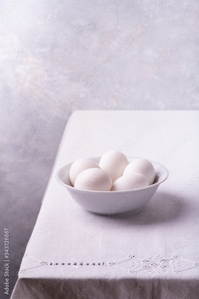 on the white linen tablecloth, in the foreground, a ceramic bowl with fresh white eggs ready for the preparation of homemade culinary recipes on the white linen tablecloth, in the foreground, a ceramic bowl with fresh white eggs ready for the preparation of homemade culinary recipes