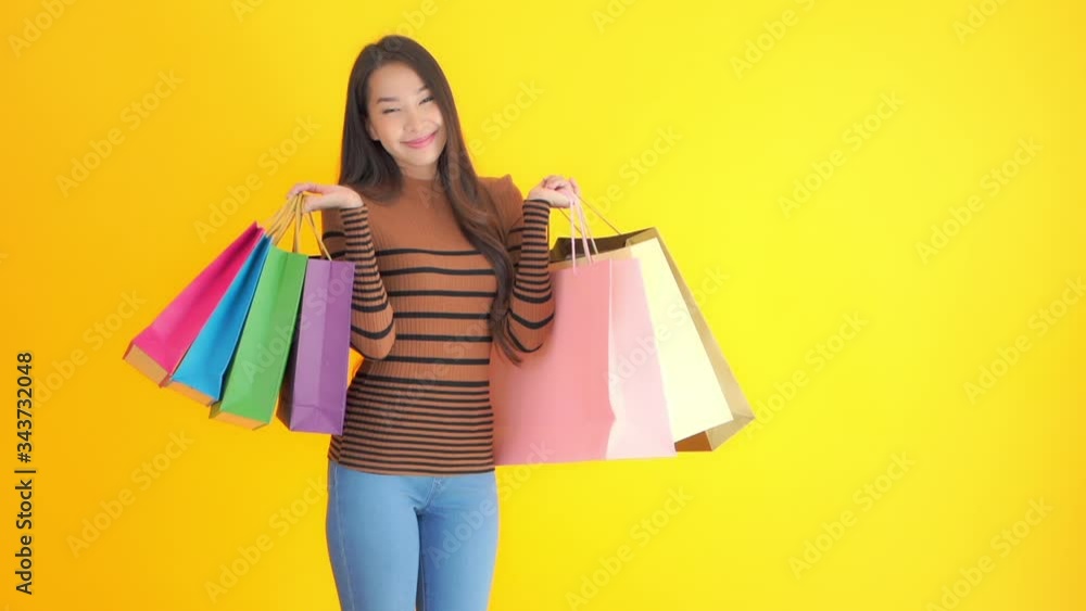 Slow-motion of Attractive Asian girl holding many shopping bags posing on yellow studio background, exited face expression Slow-motion of Attractive Asian girl holding many shopping bags posing on yellow studio background, exited face expression