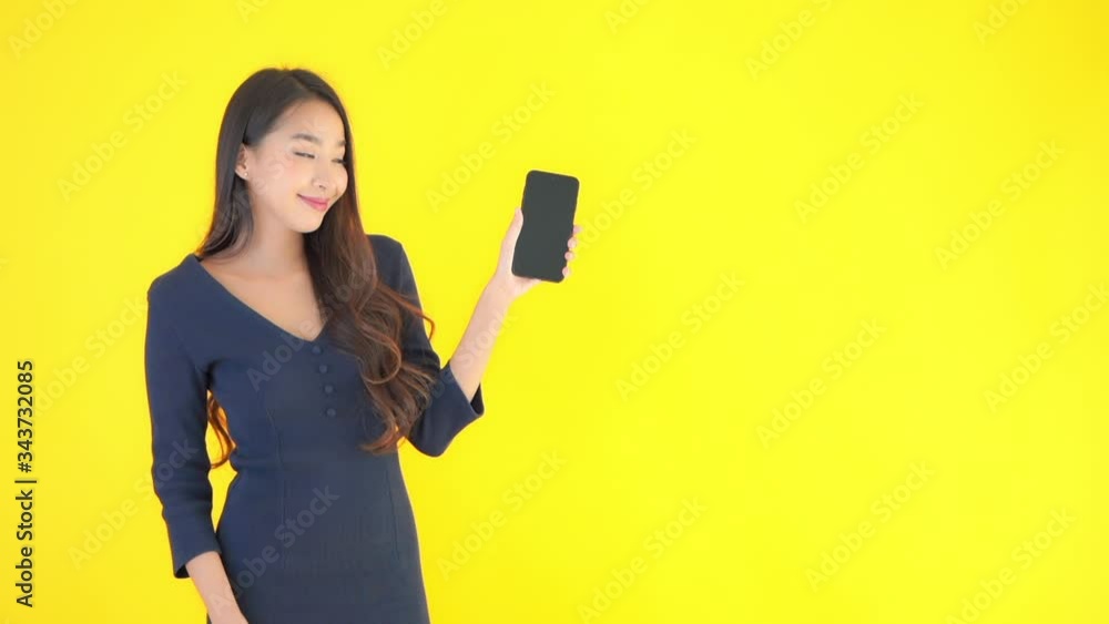 Asian girl showing phone and pointing with her finger on a black blank screen on yellow studio background Asian girl showing phone and pointing with her finger on a black blank screen on yellow studio background
