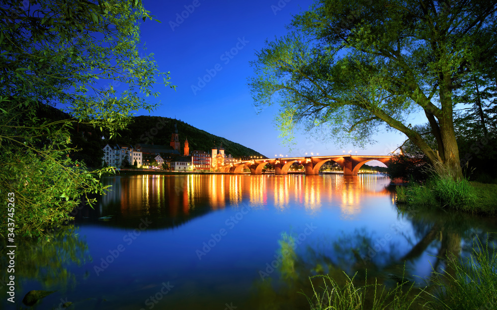 Neckar river in Heidelberg, Germany, at dusk, with deep blue sky reflected in the clear water and green trees framing the illuminated Old Bridge