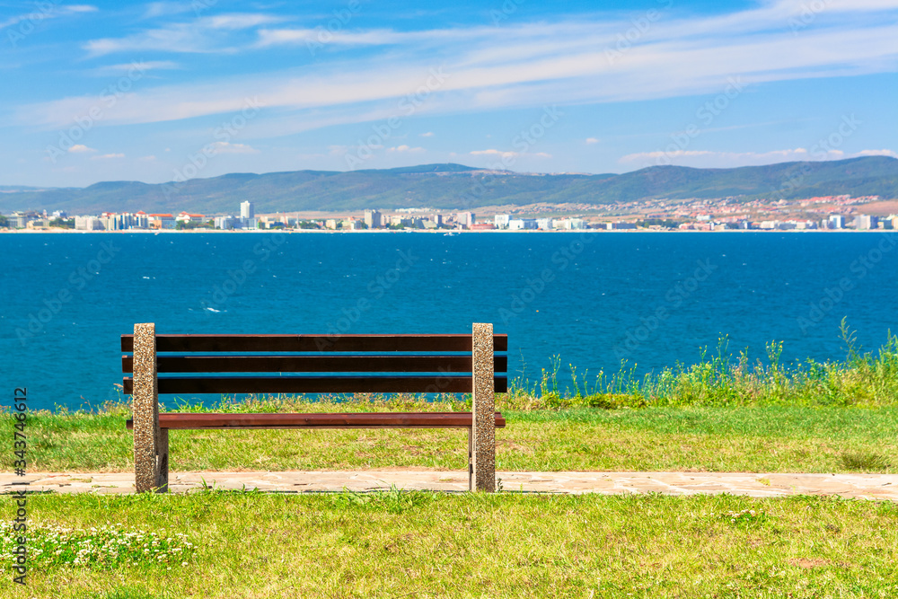 bench on the sunny beach shore. beautiful skyline view from empty park with paved footpath on the seaside. city and mountain in the distance beneath a blue sky with clouds