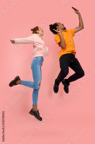 Full length side profile body size photo funny crazy people red haired female and African American guy jump high show v-sign make take selfies wear casual jeans orange tshirts isolated pink background