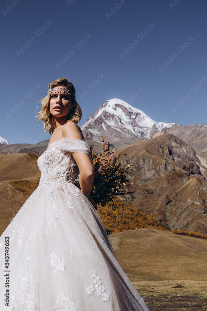 young and attractive woman dressed in wedding dress in boho style posing for photo on mountains background, perfect makeup, hairstyle and wedding floristry, happy bride in anticipation of groom