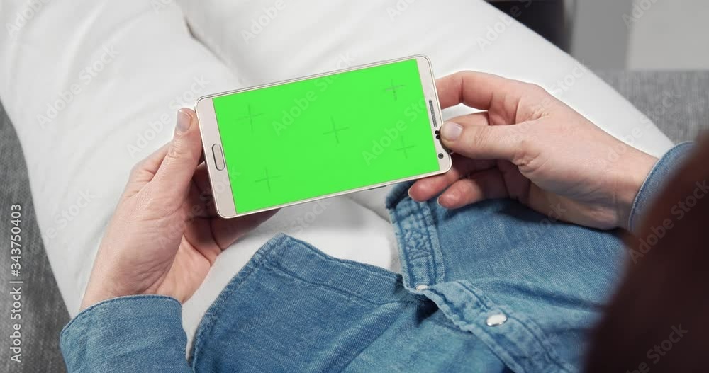 Back view of woman with brown hair lying comfortably on sofa and holding smartphone with chroma key screen. Young lady spending leisure time with device at home. Back view of woman with brown hair lying comfortably on sofa and holding smartphone with chroma key screen. Young lady spending leisure time with device at home.